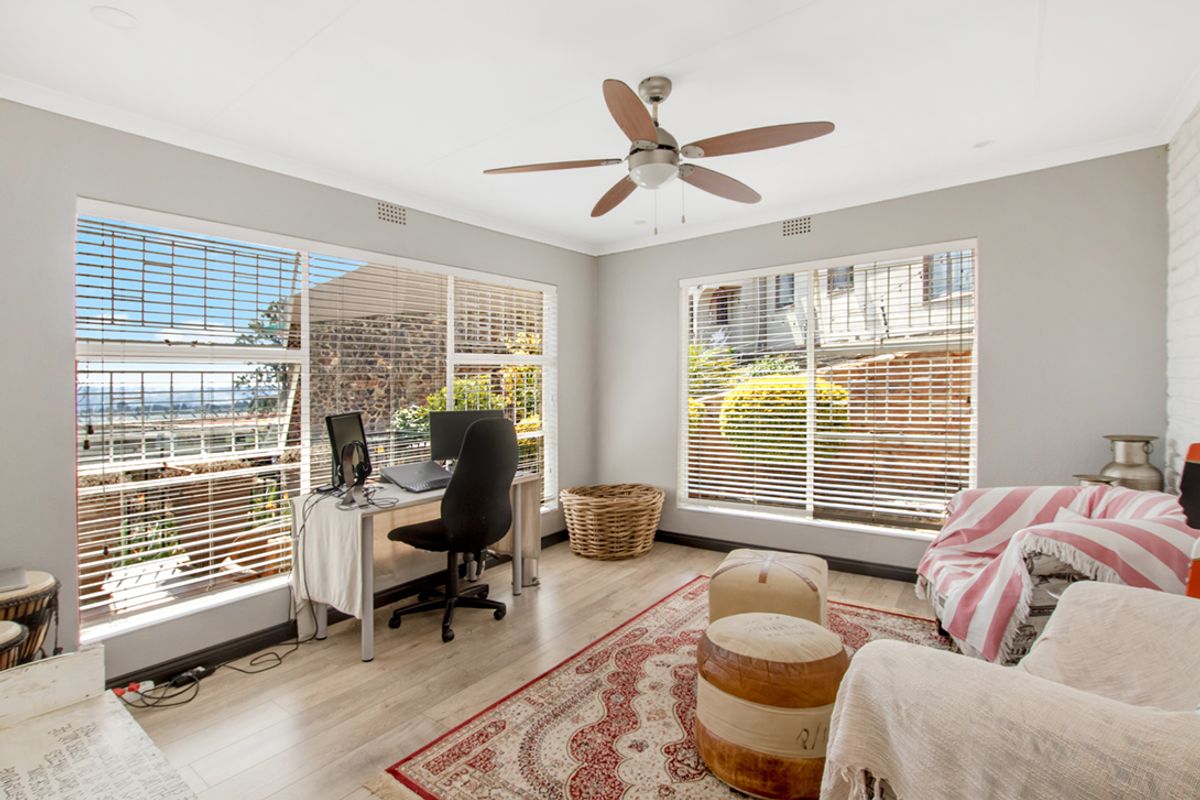 Bright living area featuring laminate flooring, a ceiling fan, and large windows with venetian blinds, offering a versatile space for relaxation or a home office.