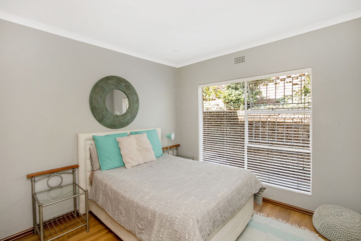 A well-lit bedroom featuring laminate flooring, neutral wall tones, and a large window with fitted venetian blinds overlooking the exterior area.
