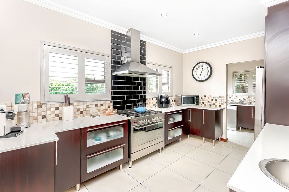 Modern kitchen featuring dark cabinetry, stainless steel gas stove with extractor fan, tiled backsplash, and light-colored floor tiles, with an adjacent scullery area.