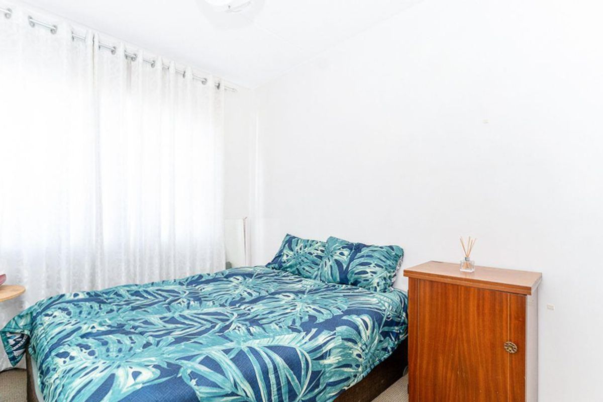 A bright bedroom featuring neutral walls, carpeted flooring, and a large window with sheer curtains, providing a simple and functional living space.