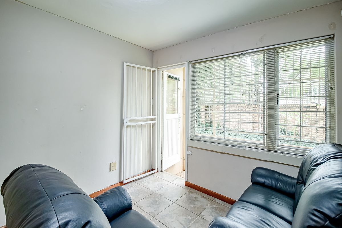 A living area featuring tiled flooring, security gate access, and large windows with venetian blinds, requiring minor cosmetic updates to the walls.