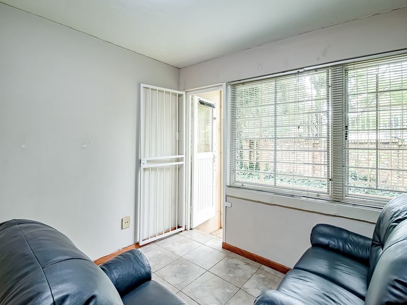 A living area featuring tiled flooring, security gate access, and large windows with venetian blinds, requiring minor cosmetic updates to the walls.