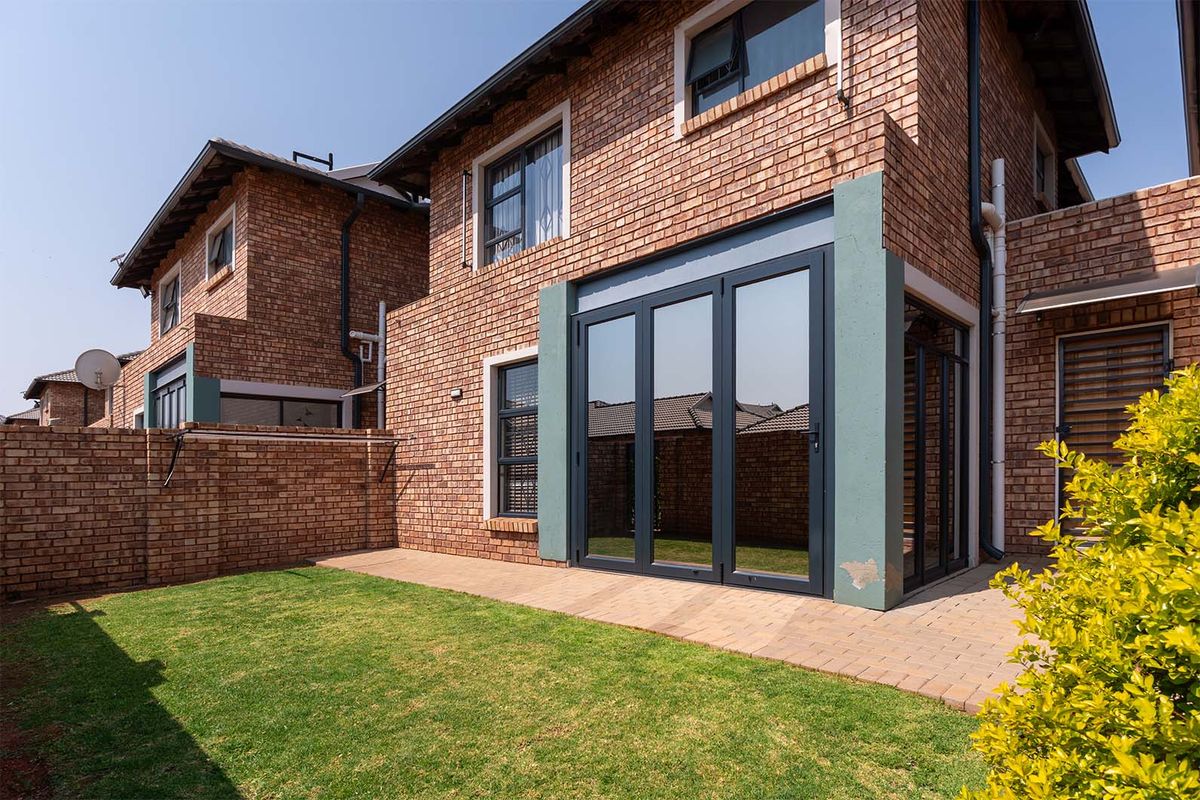 Rear view of a face-brick cluster home featuring a paved patio area, manicured lawn, and modern dark-framed folding glass doors.