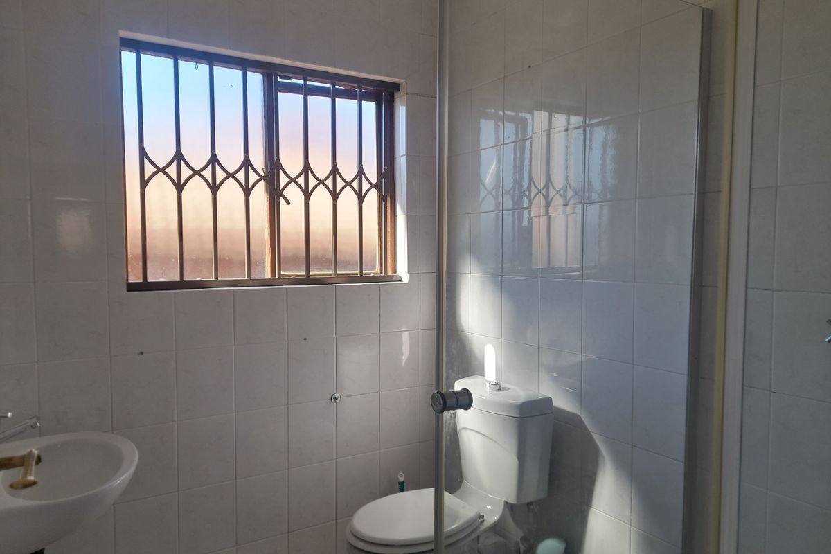 A functional bathroom featuring white wall tiles, a standard toilet, and a wall-mounted basin with security bars on the window.