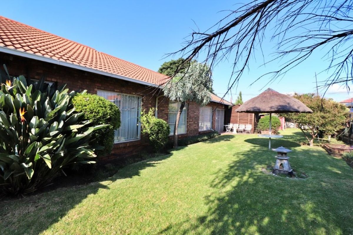 A view of the rear garden featuring a well-maintained lawn, established landscaping, and a thatched lapa area adjacent to the brick-clad house.