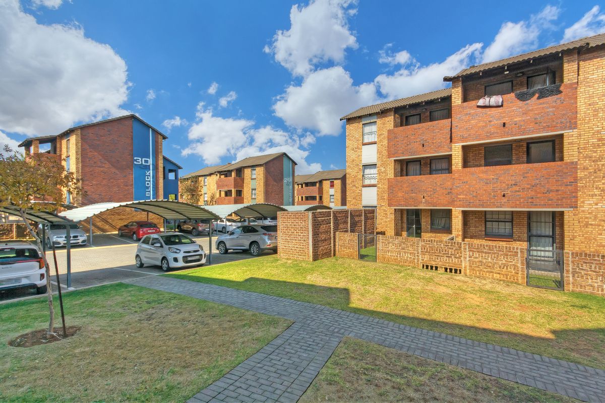 Exterior view of a multi-story brick apartment complex featuring covered parking bays, paved walkways, and communal garden areas.