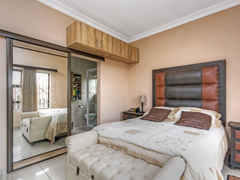 A well-maintained bedroom featuring built-in sliding mirror wardrobes, overhead storage cabinetry, tiled flooring, and direct access to an en-suite bathroom.