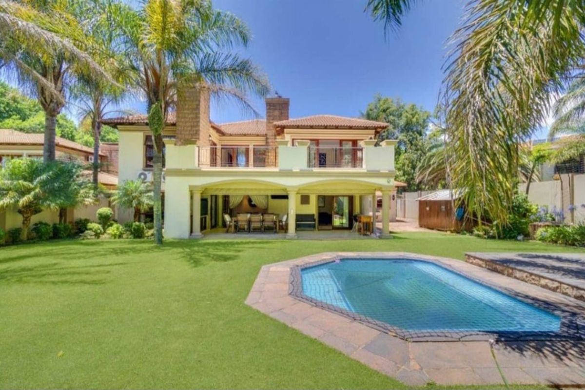 Rear view of the property featuring a spacious covered patio, upper balcony, swimming pool, and a well-maintained landscaped garden.