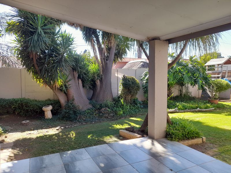 A covered patio area featuring tiled flooring and a concrete support pillar, overlooking a landscaped garden with mature trees and a boundary wall.