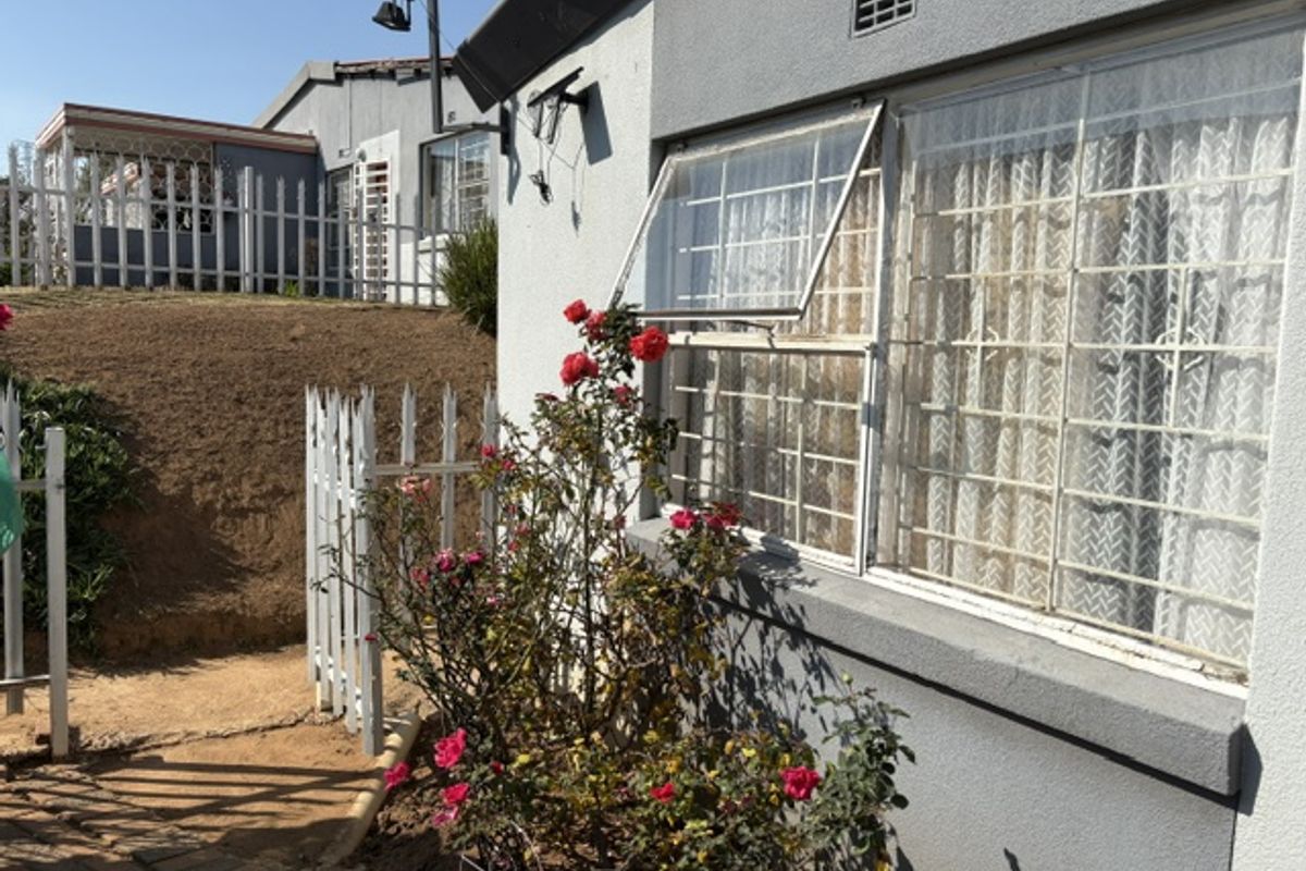 Rear exterior view of the apartment featuring a paved walkway, security-barred windows, and a white picket fence bordering an elevated garden area.