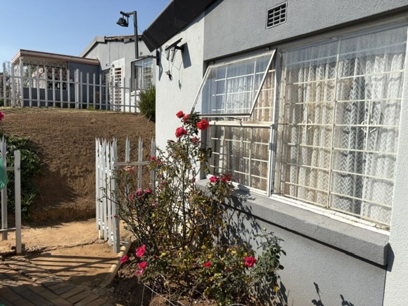 Rear exterior view of the apartment featuring a paved walkway, security-barred windows, and a white picket fence bordering an elevated garden area.
