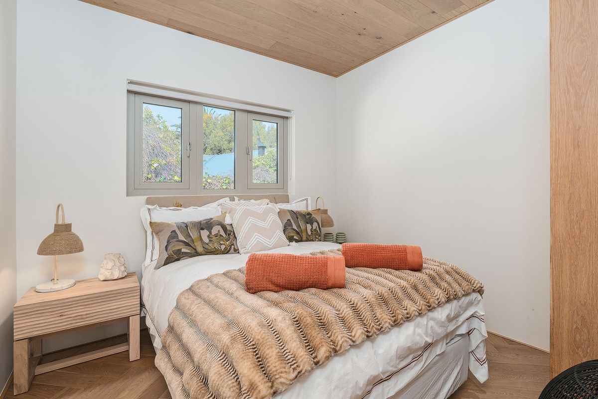A modern bedroom featuring light timber ceiling paneling, herringbone wood flooring, and a neutral color palette with a window providing natural light.