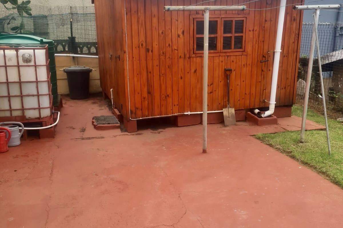 A detached timber garden shed or utility structure featuring a corrugated metal roof, situated on a paved area with an adjacent water storage tank.