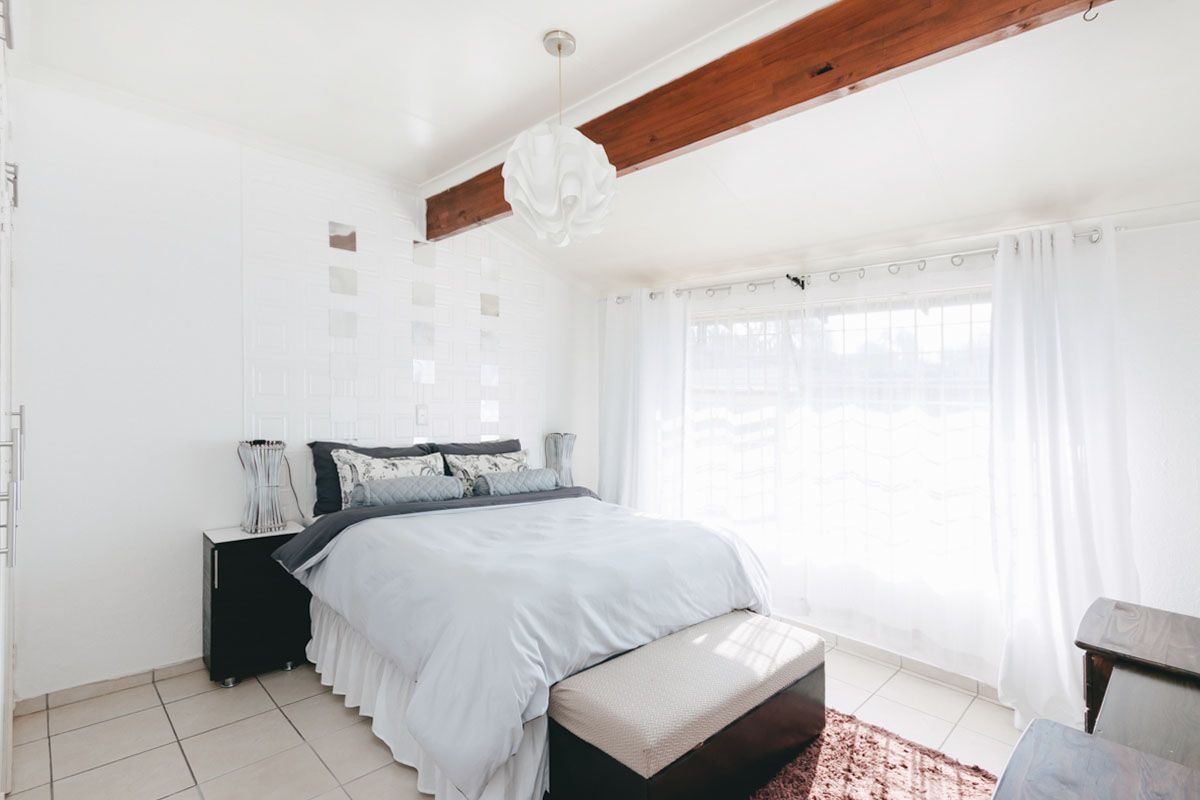 A bright bedroom featuring exposed timber ceiling beams, tiled flooring, and a textured feature wall with integrated mirror accents.
