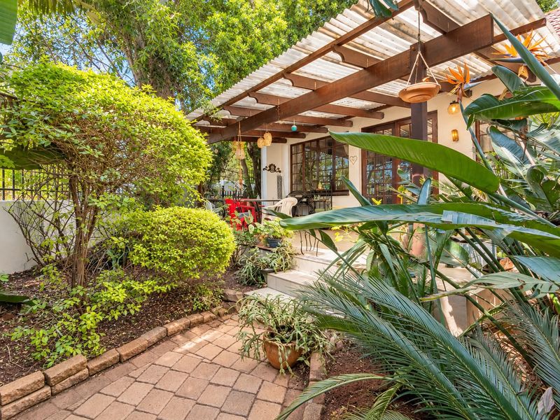 A covered outdoor patio area featuring a timber pergola with corrugated roofing, accessible via concrete steps from a brick-paved garden path.