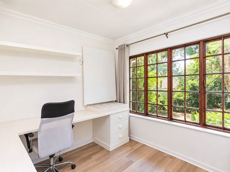 A bright home office featuring a built-in desk, integrated shelving, and large timber-framed windows overlooking a lush garden.