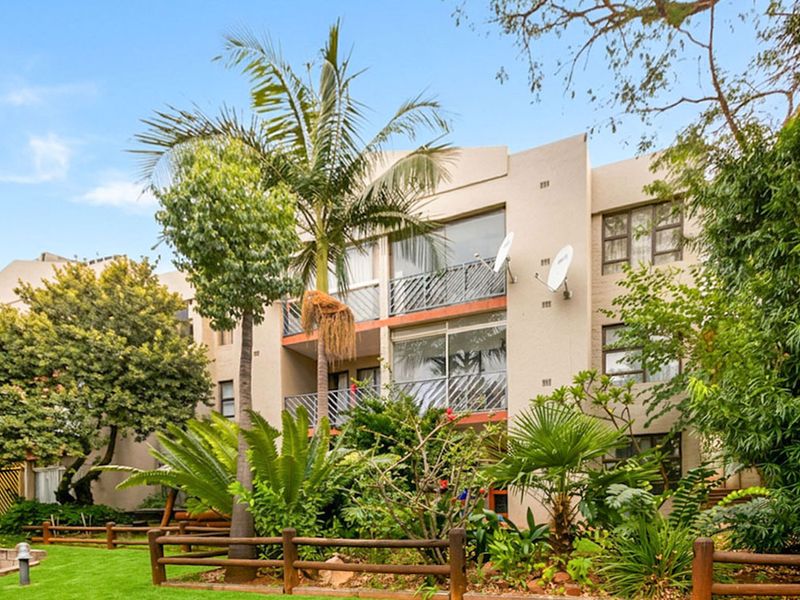 Rear elevation of a multi-story apartment building featuring balconies with metal railings, sliding glass doors, and established garden landscaping.