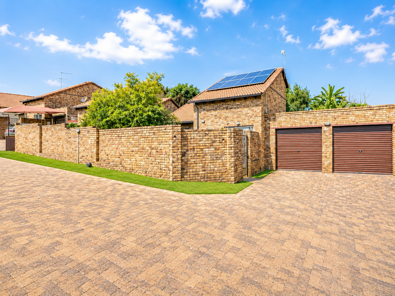 A well-maintained face-brick townhouse exterior featuring a double garage with roller doors, solar panels on the roof, and a paved driveway.