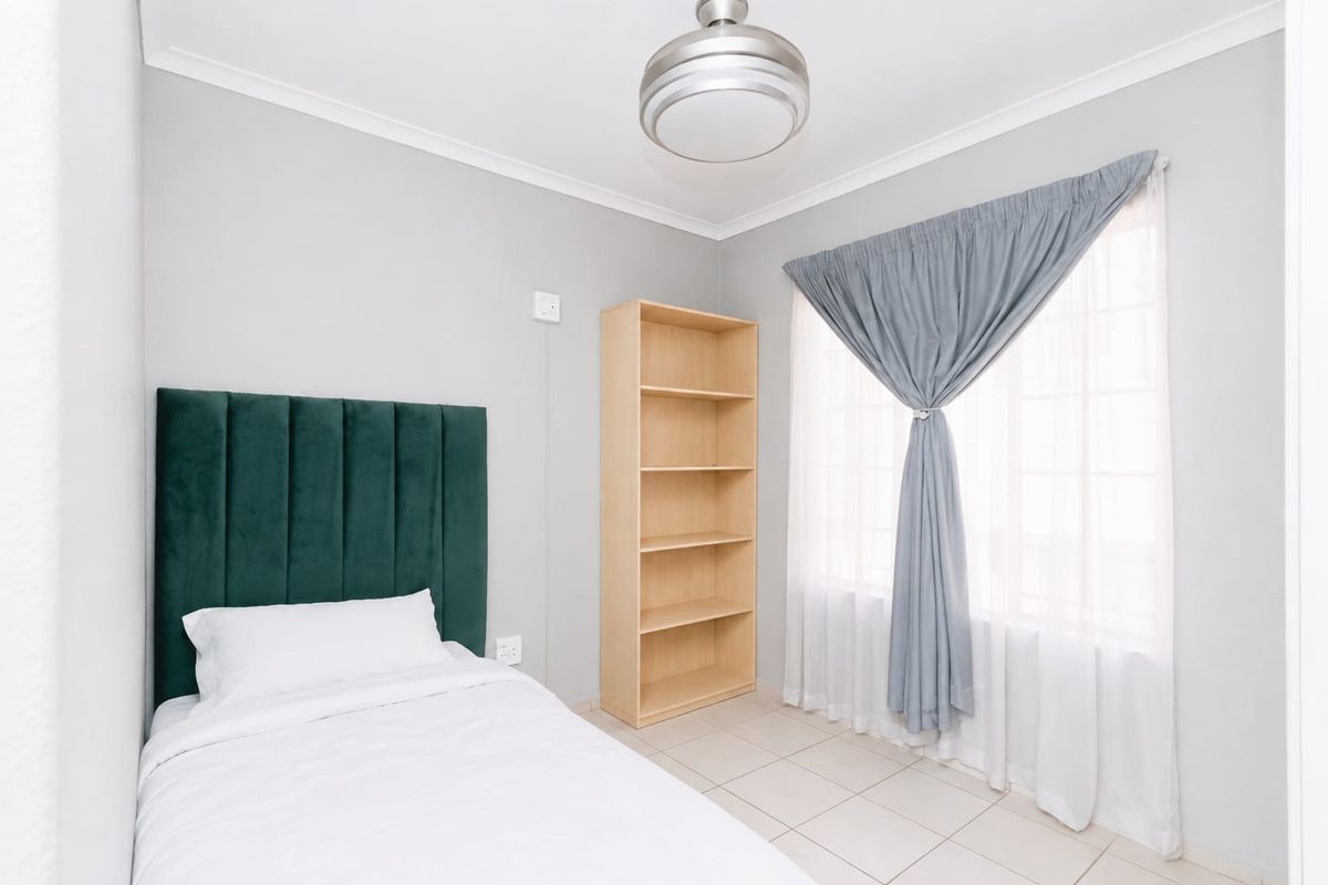 A bright bedroom featuring neutral wall tones, tiled flooring, a large window with curtains, and a built-in shelving unit.