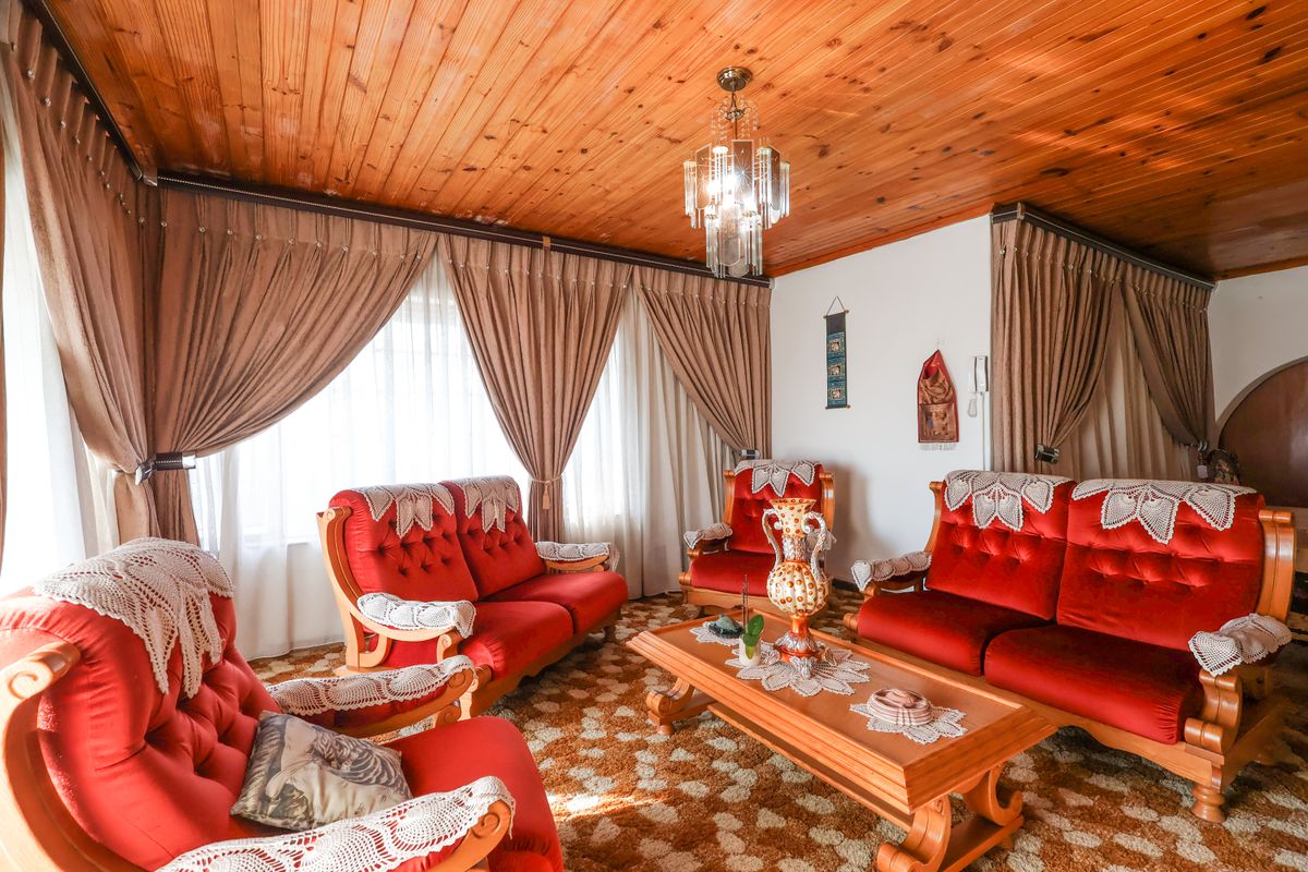 A traditional living room featuring a timber tongue and groove ceiling, a central crystal chandelier, and large windows with full-length curtains.