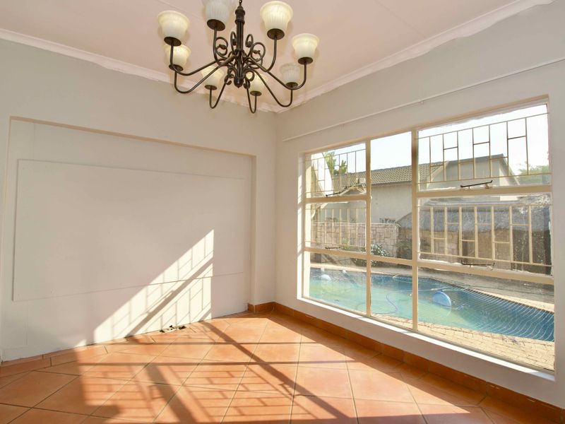 A bright, empty living room featuring terracotta floor tiles, a decorative chandelier, and a large window overlooking the swimming pool area.