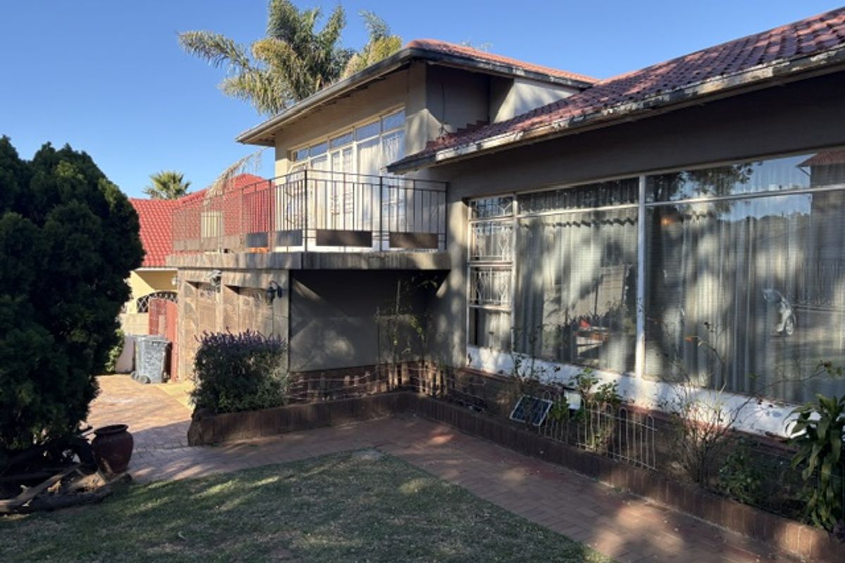 Rear view of the house featuring a raised balcony with metal railing, large windows, and a paved garden area with brick retaining walls.