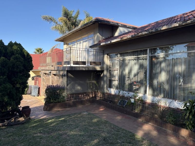 Rear view of the house featuring a raised balcony with metal railing, large windows, and a paved garden area with brick retaining walls.