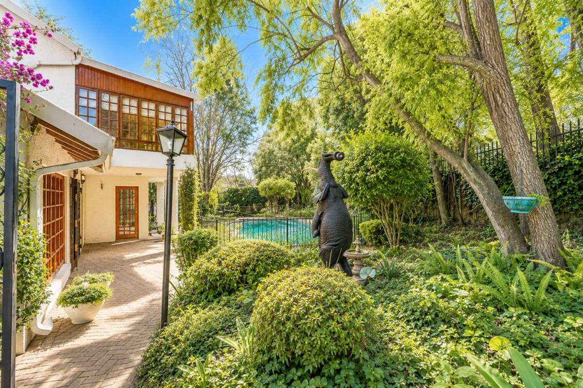 A lush rear garden featuring a brick-paved patio, mature landscaping, and a swimming pool enclosed by wrought iron fencing.