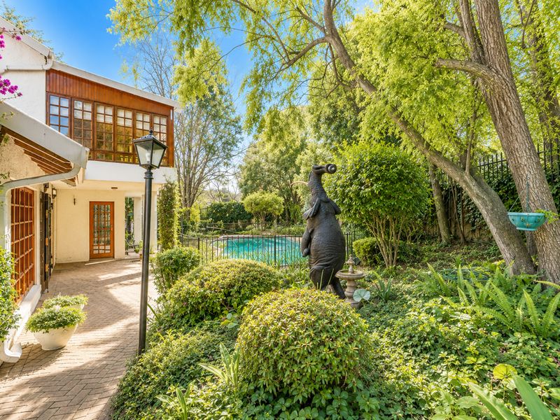 A lush rear garden featuring a brick-paved patio, mature landscaping, and a swimming pool enclosed by wrought iron fencing.
