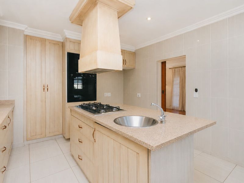 A clean, functional kitchen featuring light wood cabinetry, stone countertops, a central island with a gas cooktop and sink, and neutral tiled finishes.