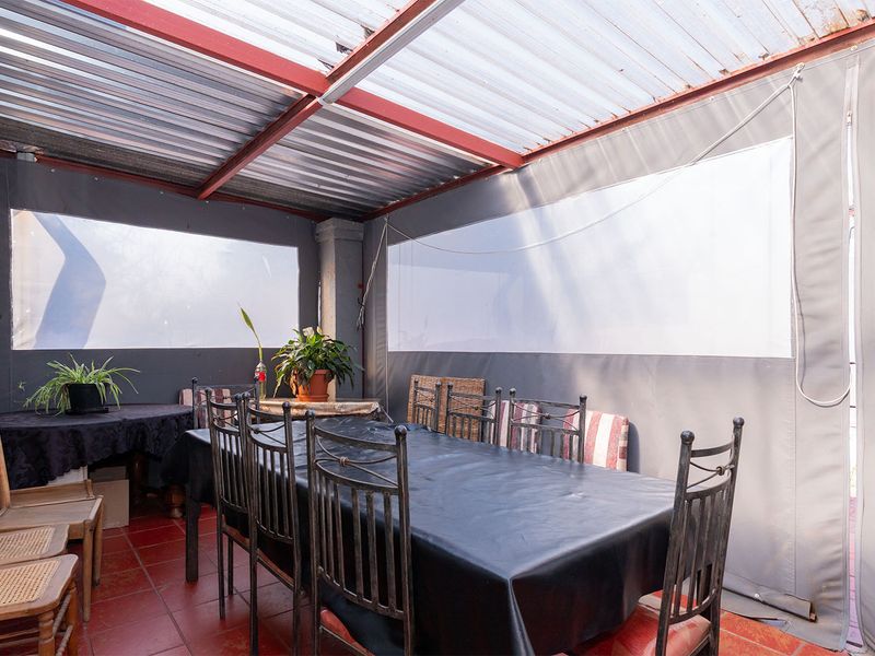 A covered outdoor patio area featuring tiled flooring, a corrugated roof structure, and retractable canvas side screens for weather protection.
