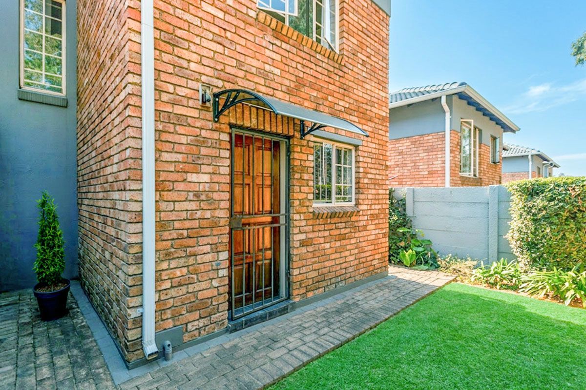 Rear exterior view of the townhouse featuring a face brick facade, security gate, protective awning, paved walkway, and a private garden area.