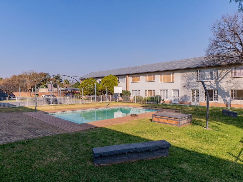 Communal outdoor area featuring a swimming pool, paved surround, and lawn, set against the apartment building facade with perimeter fencing.