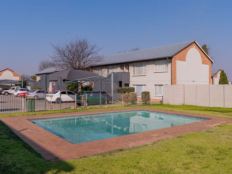 Communal swimming pool area featuring brick coping, set within a complex with established lawn, perimeter fencing, and adjacent carport parking facilities.