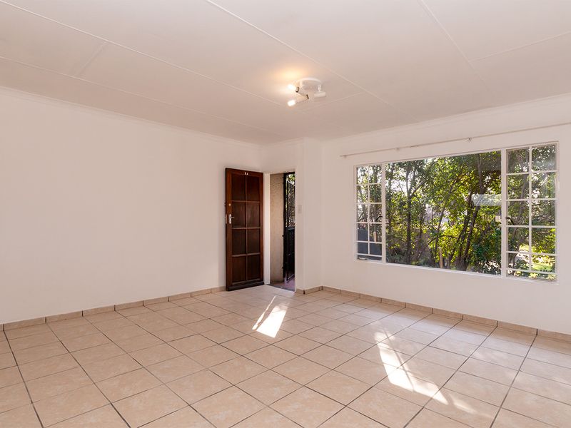 A bright, empty living room featuring neutral tiled flooring, a large window with garden views, and a classic timber entrance door.
