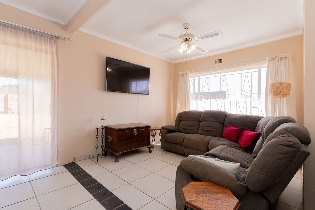 A bright living room featuring tiled flooring, a ceiling fan, and large windows with security bars, providing access to the outdoor area.