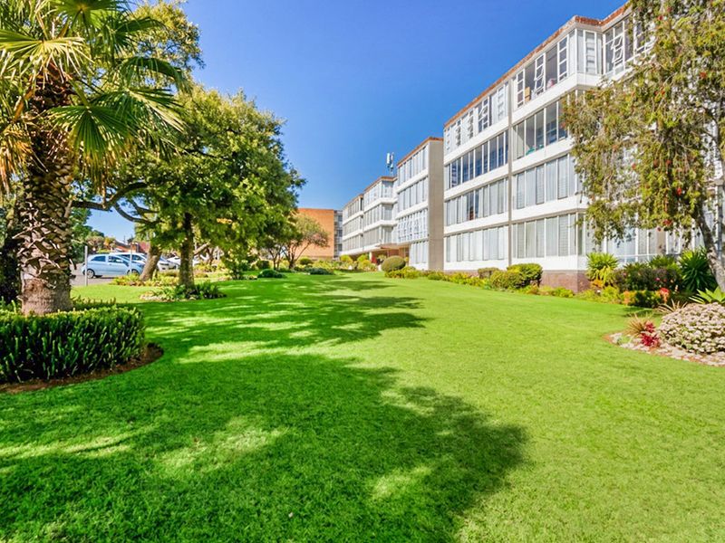 A well-maintained apartment building exterior featuring expansive green lawns, mature landscaping, and multiple levels of enclosed balconies.