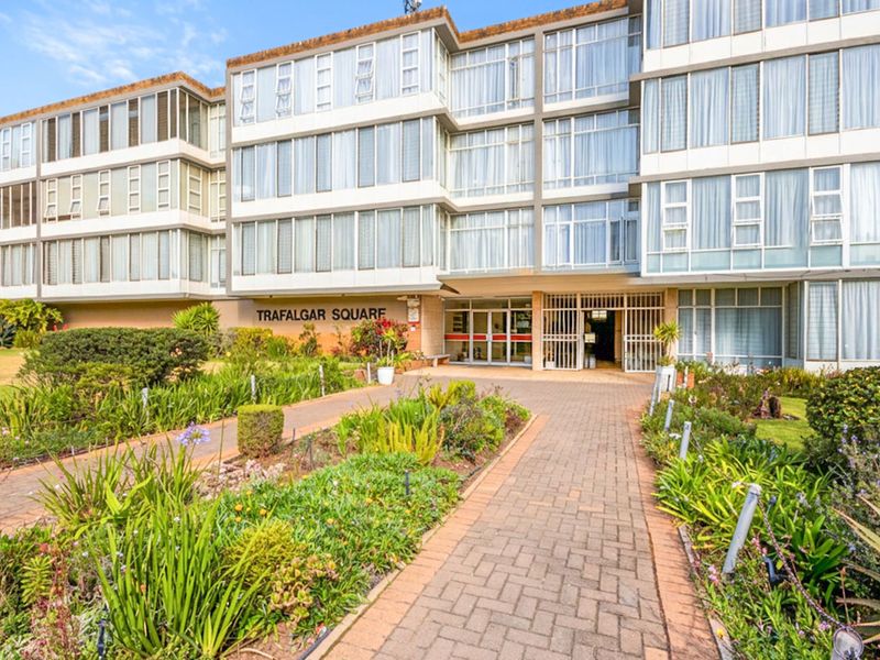 Front exterior view of Trafalgar Square apartment complex featuring a paved entrance walkway, landscaped gardens, and a multi-story building facade with large windows.