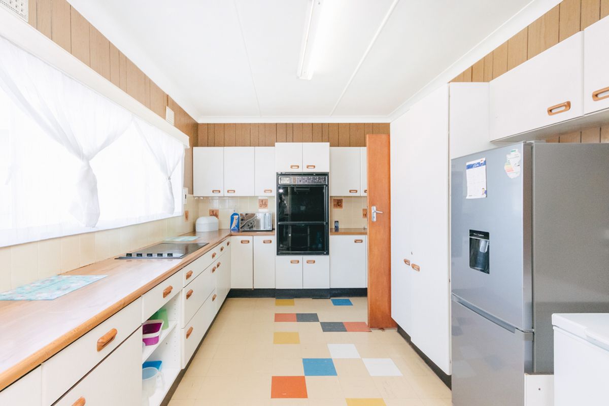 A dated kitchen featuring white cabinetry, built-in oven, cooktop, and laminate countertops with wood-look wall paneling and multi-colored vinyl flooring.