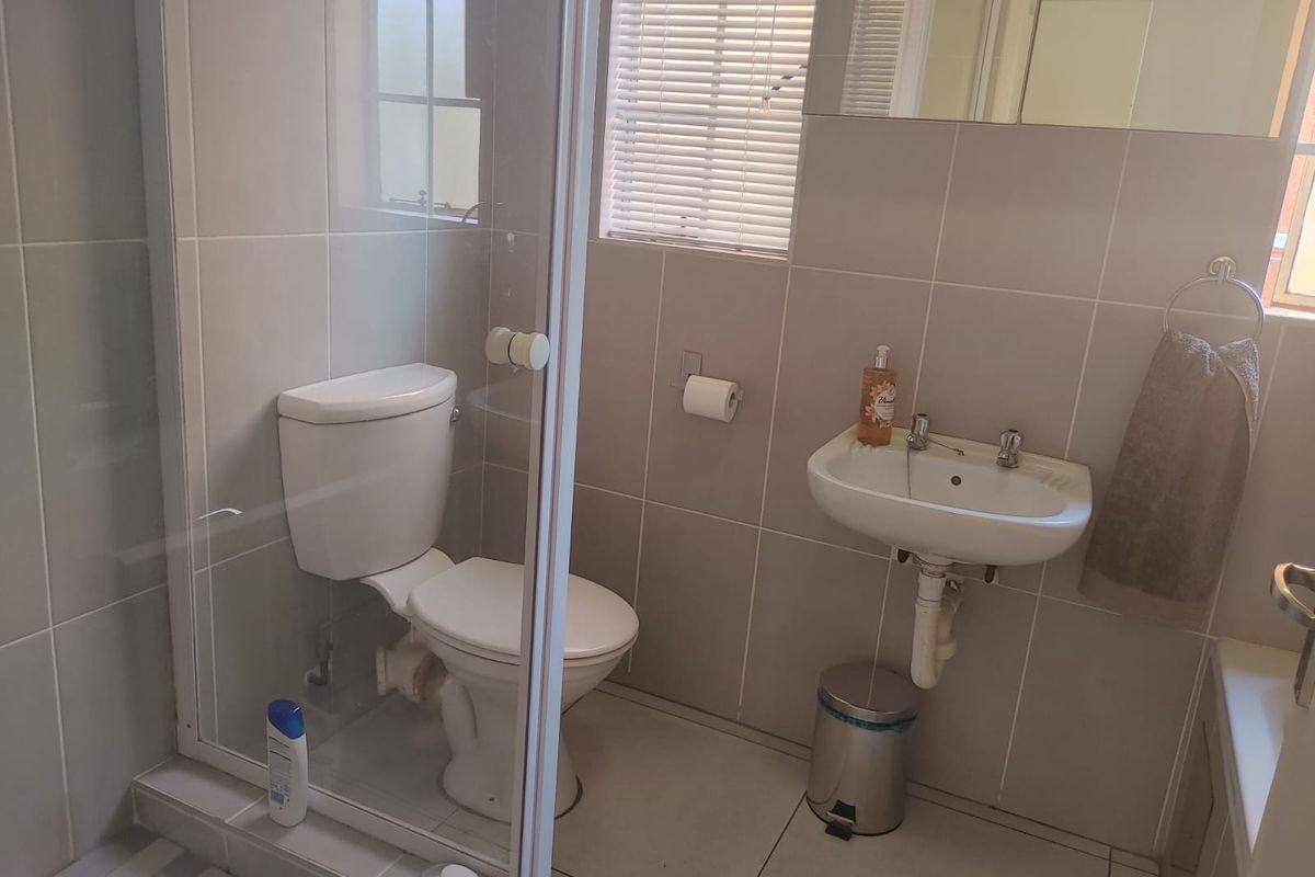A functional bathroom featuring a glass-enclosed shower, wall-mounted basin, and neutral wall tiling with natural light provided by a window with venetian blinds.