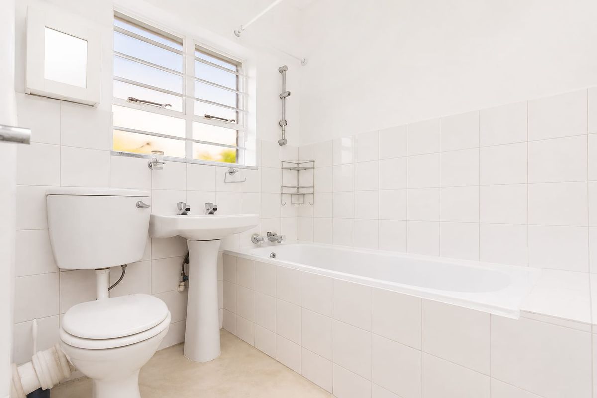 A clean, functional bathroom featuring a white pedestal basin, toilet, and bathtub with tiled walls and a louvered window for natural ventilation.