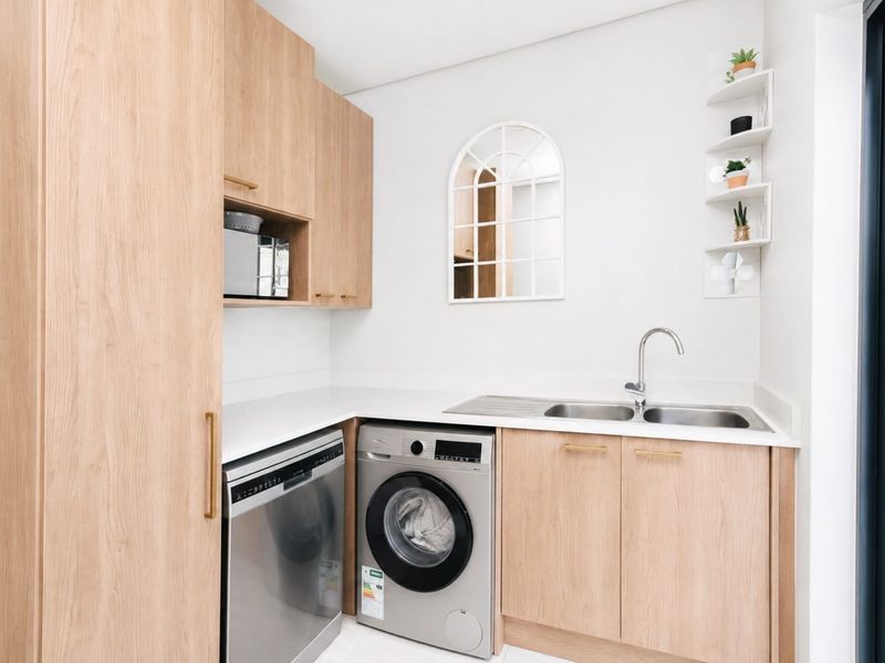 Modern laundry room featuring light wood cabinetry, stone countertops, a stainless steel double sink, and dedicated space for integrated appliances.