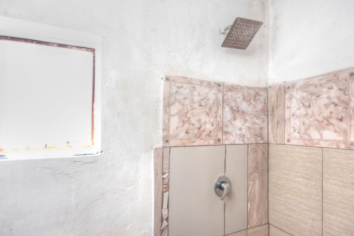A bathroom shower area featuring a fixed shower head and mixer, requiring maintenance to the wall finishes and window frame.