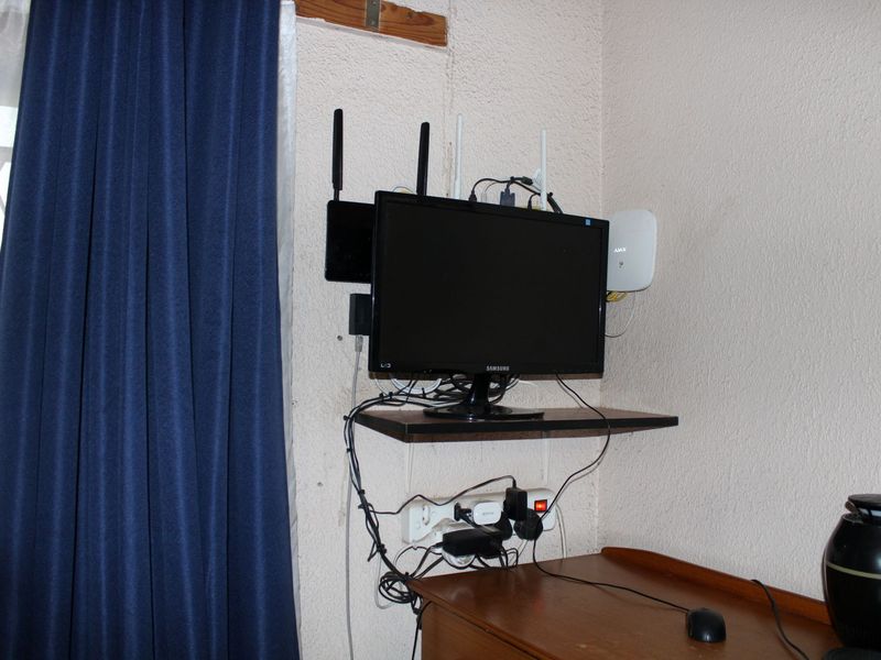 A bedroom corner featuring textured walls and a window with security bars, currently utilized as a workspace with various electronic equipment.