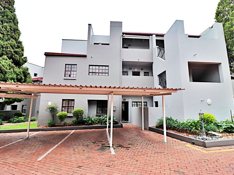 Front exterior view of a multi-story apartment complex featuring a paved driveway, covered carport parking, and established garden beds.