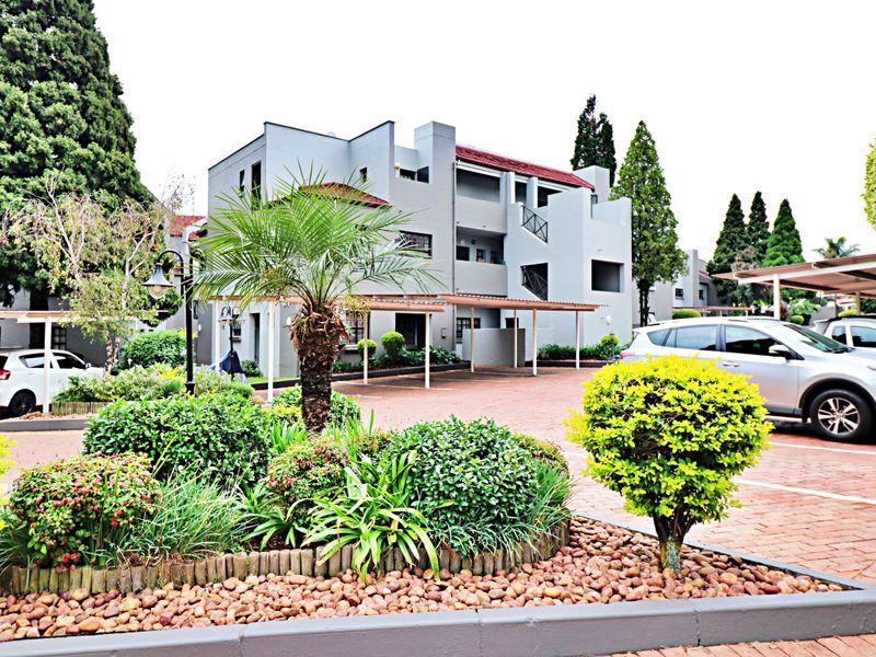 Exterior view of an apartment complex featuring a paved driveway, covered parking bays, and well-maintained landscaped garden areas.