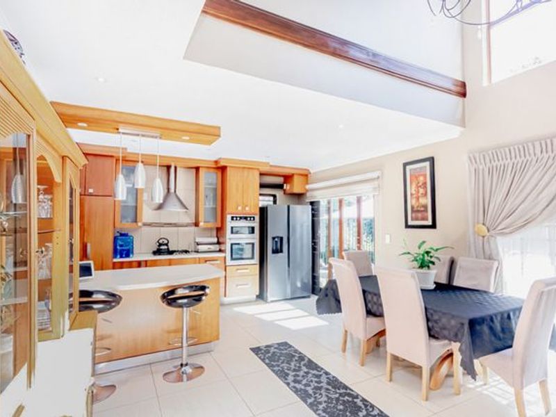 Spacious open-plan kitchen and dining area featuring wooden cabinetry, built-in appliances, tiled flooring, and a double-volume ceiling with abundant natural light.
