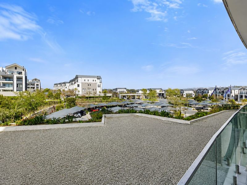 A spacious balcony featuring a modern glass balustrade and gravel surface, offering an elevated view over the residential complex and parking area.
