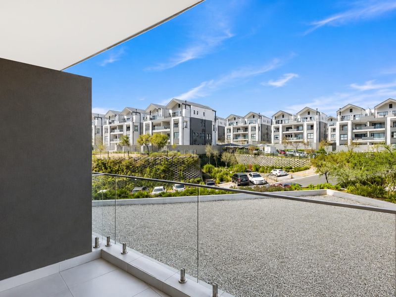 A modern balcony featuring a sleek glass balustrade and tiled flooring, offering an elevated view of the surrounding apartment complex and landscaped grounds.
