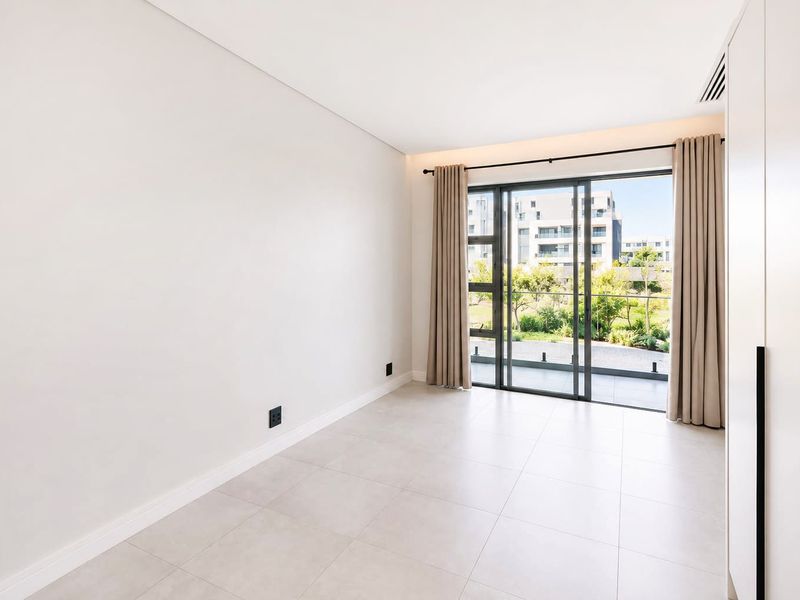 A bright, modern, and empty bedroom featuring large sliding glass doors leading to a balcony, neutral tiled floors, and built-in storage cabinetry.